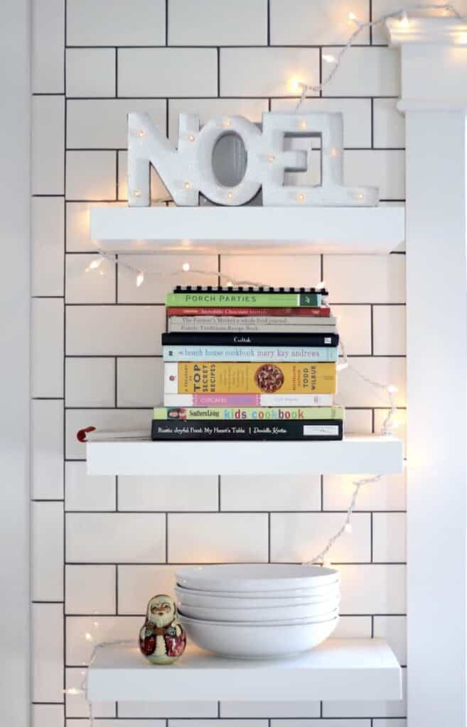 How to Host a Christmas Movie Marathon & Cookie Swap. Image of a floating shelves in a white kitchen decorated with Christmas lights and other decor.