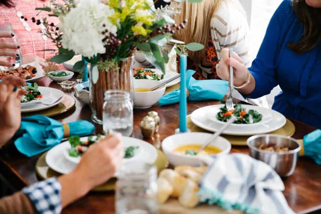 How to Host a Simple Dinner Party Like a Pro. A holiday table full of food with hands of women eating and sharing a meal together.