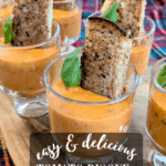 Close-up of small clear glasses filled with creamy orange-colored tomato bisque soup, garnished with a fresh basil leaf and a toasted grilled cheese sandwich crust slice.