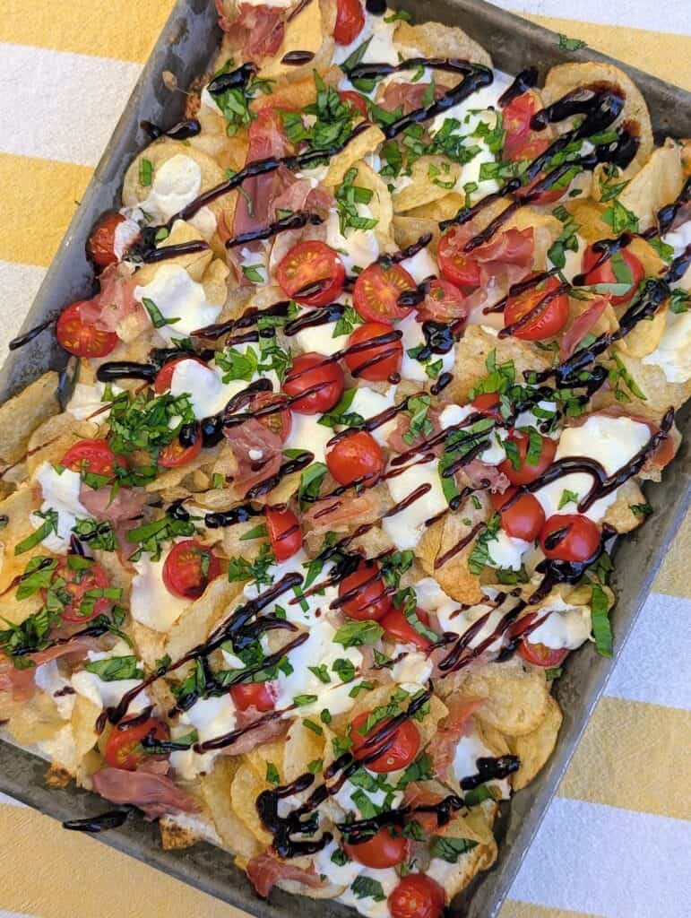 The Viral Potato Chip Charcuterie: Caprese Style of Crispy potato chips topped with cherry tomatoes, fresh basil, balsamic glaze, creamy cheese, and prosciutto, garnished with chopped herbs.