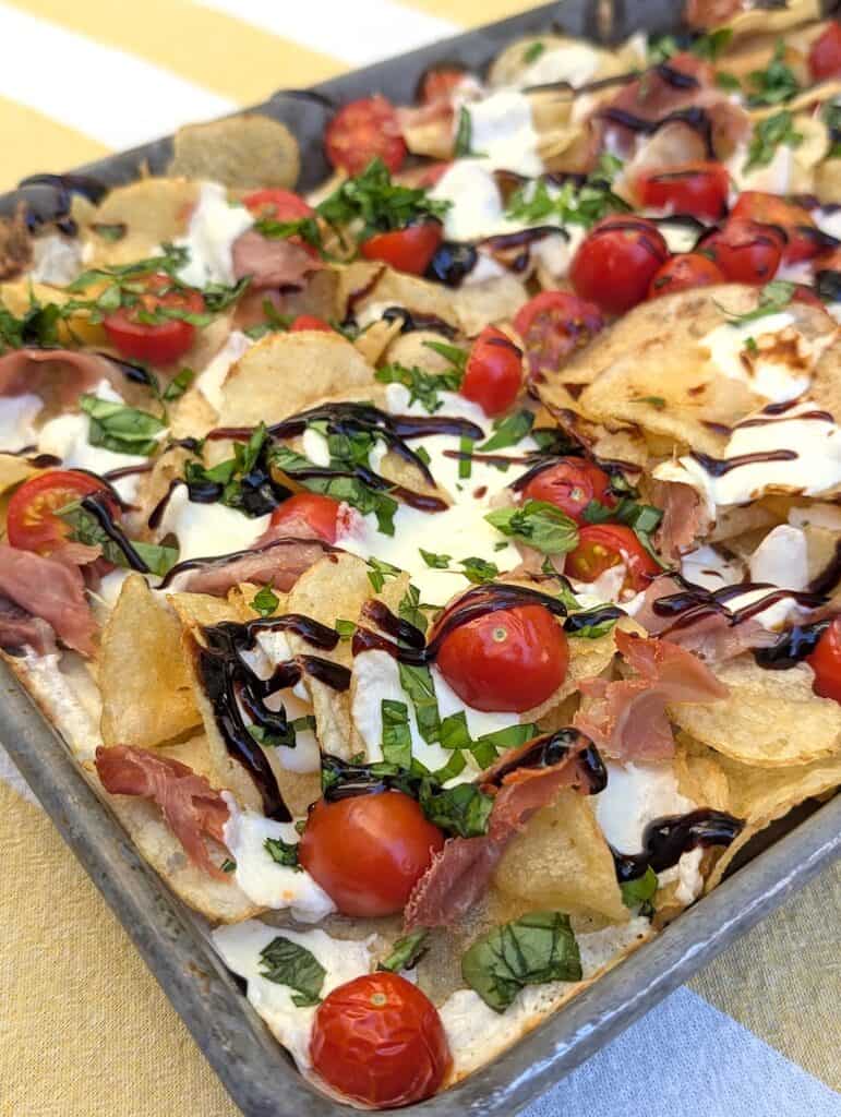 A full sheet pan of potato chips, with fresh mozzarella and cherry tomatoes scattered on top, and garnished with prosciutto, fresh basil, and a balsamic glaze. The Viral Potato Chip Charcuterie: Caprese Style