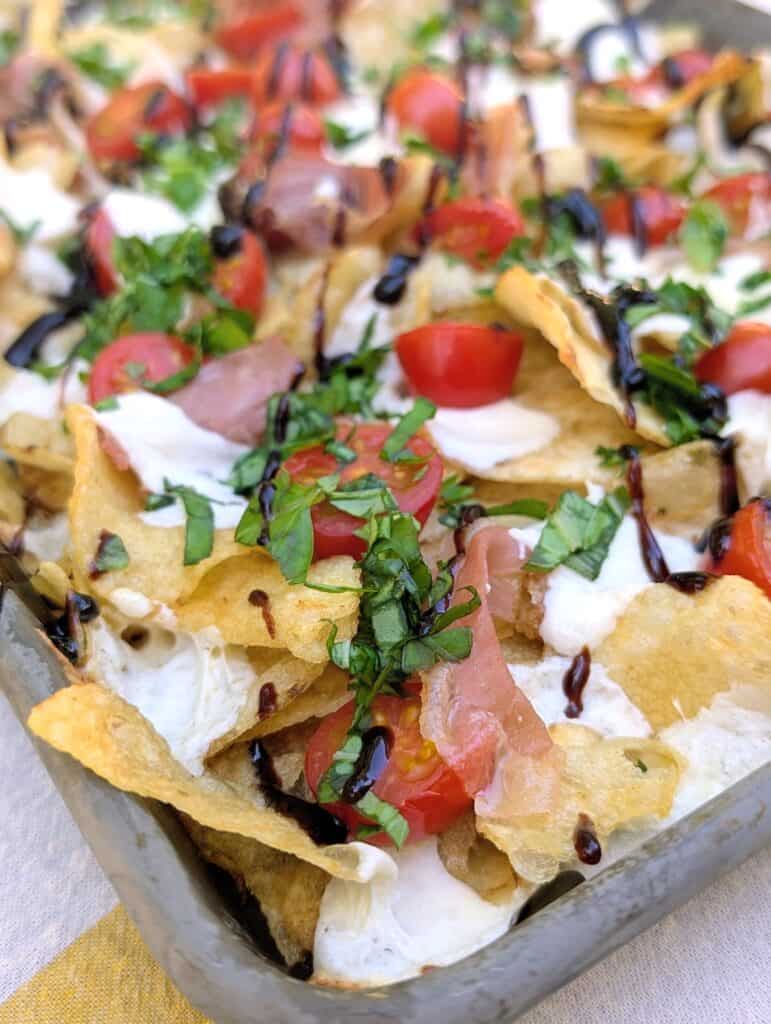 A close-up of a delicious and savory snack tray of charcuterie chips, caprese style, with crispy potato chips covered in melty mozzarella, bright red cherry tomatoes, and savory prosciutto, garnished with green herbs and a dark drizzle