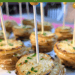 A low-angle close-up of a stack of crispy, baked potato slices (likely cooked in a muffin tin), topped with Parmesan and parsley, and held together with a tall, gold-tipped pick. Text overlay reads: "how to make garlic & parmesan Crispy Potato Stacks in muffin tins."