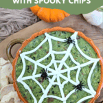 Overhead photo of a festive Halloween Guacamole Dip in a pumpkin-shaped bowl, decorated with a spider web made of white cream and plastic spiders. Spooky-shaped chips surround the dip. Text overlay reads: "easy & delicious Halloween Guacamole with Spooky Chips."