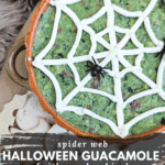Close-up of guacamole served in a pumpkin-shaped bowl, decorated with a sour cream spider web and two black plastic spiders. The bowl is surrounded by cutout, spooky-shaped tortilla chips. Text overlay reads: "spider web Halloween Guacamole {with spooky chips}."