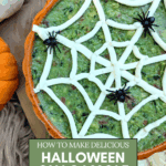 Overhead close-up of a pumpkin-shaped bowl of guacamole topped with a white cream spider web and two black plastic spiders. The bowl is next to small orange and white pumpkins. Text overlay reads: "How to Make Delicious Halloween Guacamole, The Easy Way."