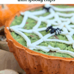 A close-up, slightly angled photo of a pumpkin-shaped dish filled with guacamole, featuring a creamy white spider web design and a plastic spider. Spooky-shaped tortilla chips are visible in the foreground. Text overlay reads: "how to make a spider web Halloween Guacamole Dip with spooky chips."