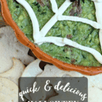Close-up of the Halloween Guacamole Dip in a pumpkin dish, showing the creamy spider web and a black plastic spider, with a pile of cutout tortilla chips in the foreground. Text overlay reads: "quick & delicious Halloween Guacamole Dip."