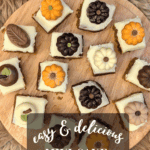 Overhead view of square pumpkin spice cake bites arranged on a round wooden cutting board. Each is topped with a white-frosted square and a chocolate in the shape of a pumpkin, leaf, or apple. Text overlay reads: "easy & delicious Mini Cake Bites Hack."