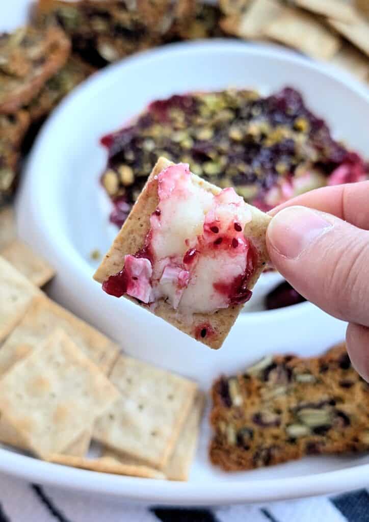 finger holding a cracked topped with baked brie with cranberry sauce