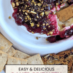 A top-down view of baked brie topped with deep red cranberry sauce and green chopped pistachios, with a serving knife partially in the cheese. Crackers are visible below. Text overlay: EASY & DELICIOUS BAKED BRIE WITH CRANBERRY SAUCE.