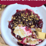 A gold knife is holding a generous scoop of melted baked brie, cranberry sauce, and pistachios over the cheese wheel. Crackers are visible on the side. Text overlay: easy & festive BAKED BRIE WITH CRANBERRY & PISTACHIOS.