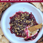 A close-up view of a baked brie wheel covered in cranberry sauce and pistachios, served with crackers. A knife is scooping out a serving. Text overlay: cozy & delicious CHRISTMAS BAKED BRIE WITH CRANBERRY.