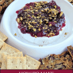 A round of warm baked brie topped generously with bright red cranberry sauce and sprinkled with chopped pistachios, served in a white dish surrounded by a variety of crackers.