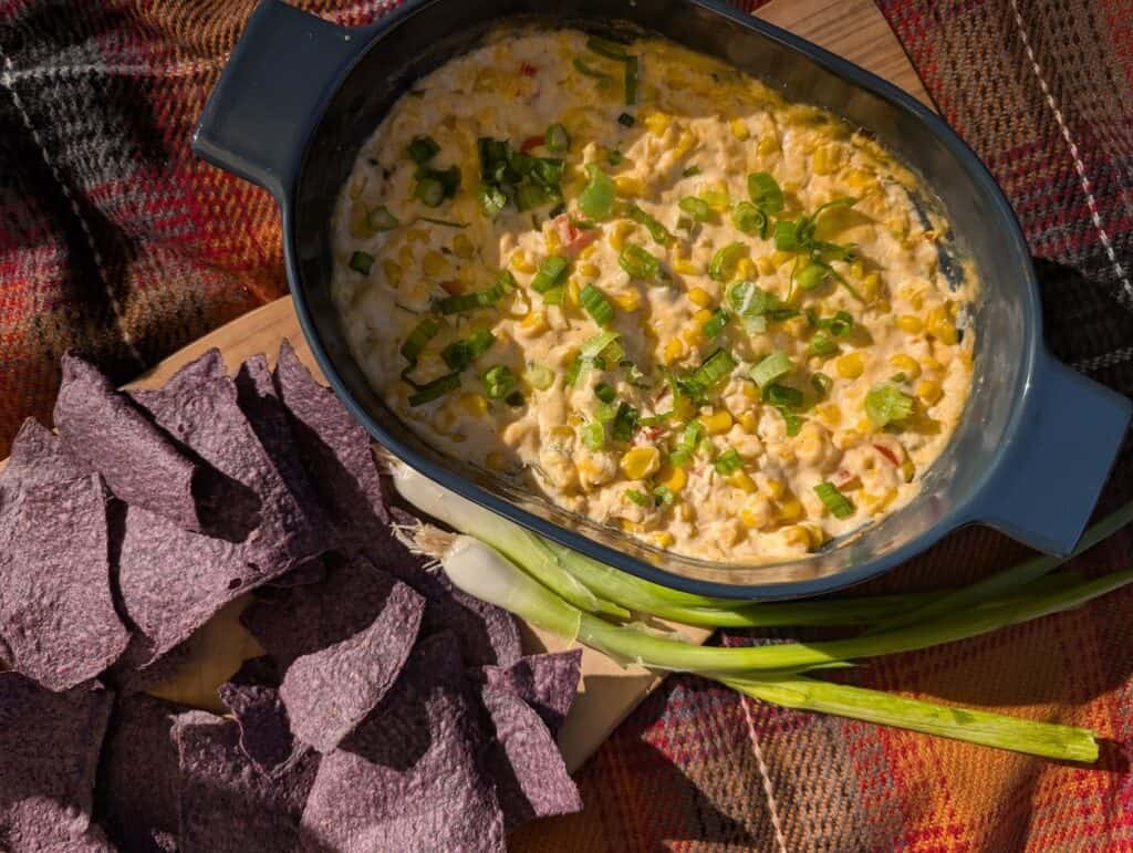 baked creamed corn dip with chips, green onion on a board on top of a plaid blanket