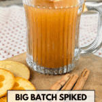 A mug of hot, spiked apple cider garnished with an apple slice, sitting on a wooden cutting board with cinnamon sticks and dried orange slices. Text overlay reads: "Big Batch Spiked Apple Cider with Fireball Whisky."