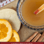 An overhead shot of a clear glass mug containing golden spiked apple cider, garnished with an apple slice. It is resting on a cutting board next to cinnamon sticks, apple slices, and orange slices. Text overlay reads: "How to Make Spiked Apple Cider with Fireball."