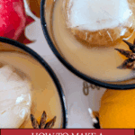 An overhead close-up of two glasses of iced, spiked apple cider cocktail. Each glass contains a large round ice cube and is garnished with a star anise. Fresh apples and oranges are visible in the background. Text overlay reads: "How to Make a Spiked Cider Cocktail with Fireball Whisky."