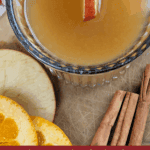 A close-up, top-down view of a clear mug filled with warm Fireball Apple Cider Hot Toddy, garnished with an apple slice. The drink is surrounded by cinnamon sticks, sliced apple, and sliced orange on a wooden cutting board. Text overlay reads: "how to make a big batch Fireball Apple Cider hot toddy."