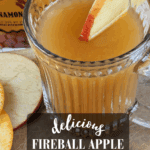 A close-up shot of a clear, ribbed mug of Fireball Apple Cider Hot Toddy, garnished with an apple wedge. A bottle of Fireball whisky is blurred in the background, along with cinnamon sticks and fruit slices. Text overlay reads: "Delicious Fireball Apple Cider Hot Toddy, a big batch recipe."
