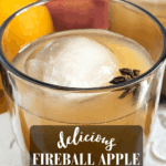 A glass of iced Fireball Apple Cider Cocktail with a large spherical ice cube and a star anise garnish. Fresh apples and an orange are visible behind the glass. Text overlay reads: "Delicious Fireball Apple Cider Cocktail, a big batch recipe."