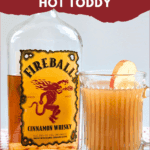 A large bottle of Fireball Cinnamon Whisky next to a mug of hot, spiked apple cider garnished with an apple slice. Dried orange slices and cinnamon sticks are scattered on the wooden board. Text overlay reads: "easy & delicious Fireball Apple Cider Hot Toddy."