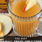 A clear, ribbed mug of spiked apple cider with an apple slice garnish, placed next to a bottle of Fireball Cinnamon Whisky. Cinnamon sticks and fruit slices are on the cutting board. Text overlay reads: "The Best Fireball Spiked Apple Cider {Big Batch Recipe}."