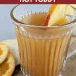 A close-up of a hot mug of Fireball Apple Cider, garnished with a crisp apple wedge. Cinnamon sticks and orange slices are visible on the wooden surface beneath the mug. Text overlay reads: "big batch Fireball Apple Cider Hot Toddy."