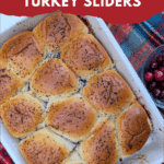 Baked cranberry turkey sliders in a white dish on a plaid surface, with a small bowl of fresh cranberries. The golden rolls are shiny and topped with poppy seeds. The text on the image reads "easy & delicious HOLIDAY CRANBERRY TURKEY SLIDERS."