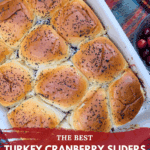 Overhead shot of a white casserole dish filled with baked Turkey Cranberry Sliders with a poppy seed topping, sitting on a red and green plaid blanket. A small bowl of fresh cranberries is visible on the side. The text on the image reads "THE BEST TURKEY CRANBERRY SLIDERS {PERFECT FOR LEFTOVERS}."