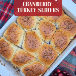 Overhead view of a baked batch of poppy seed topped cranberry turkey sliders in a white baking dish, with a small bowl of fresh cranberries to the side on a plaid blanket. The text on the image reads "easy & delicious CRANBERRY TURKEY SLIDERS."