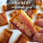 A hand holding one candied bacon cracker, showing the glossy maple-glazed bacon strip on top of the cracker. Other crackers are blurred in the background. The text overlay reads: "delicious CANDIED BACON CRACKERS with maple syrup."