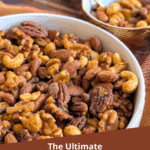 The Ultimate Candied Spiced Nuts with maple syrup and chili crisp. A large white bowl of glazed mixed nuts sits on a cozy plaid blanket, with a small gold bowl of nuts in the background.