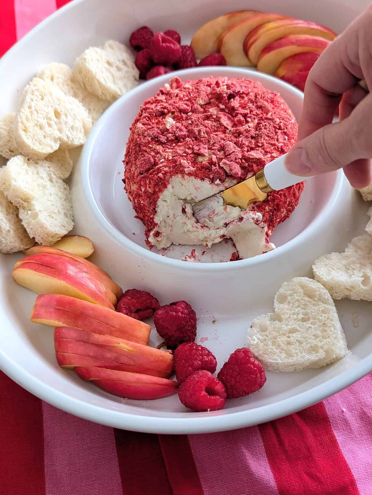 Hand slicing into a strawberry cheese ball coated in freeze-dried strawberries on a white serving platter.