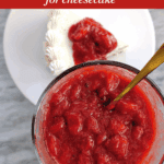 Hand holding bowl of strawberry sauce with spoon and cheesecake slice in background.
